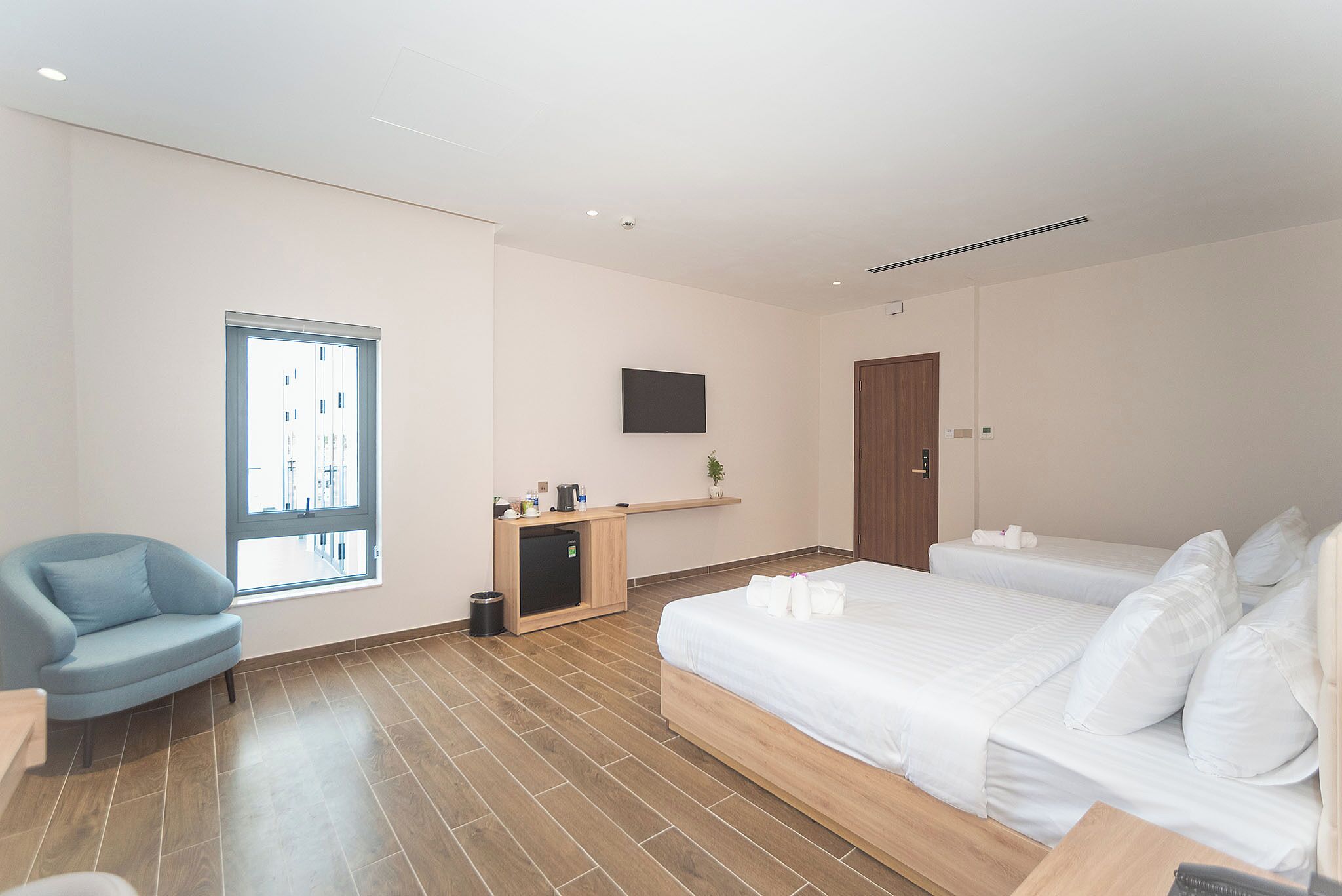 A modern hotel room with a large bed, a desk, a chair, a small kitchenette, and a TV mounted on the wall.