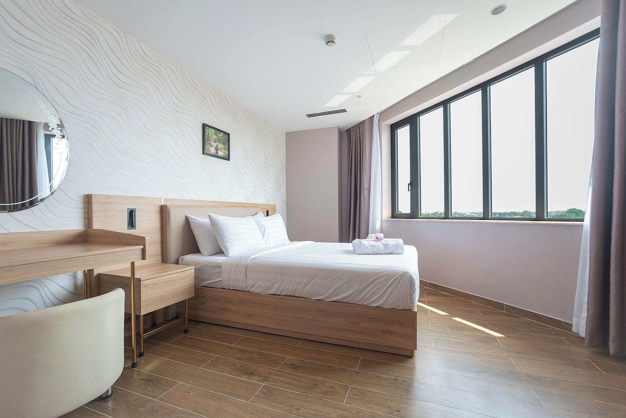 Moderni hotellihuone, jossa on puinen sänky, yöpöytä, suuri peili ja suuri ikkuna, josta avautuu näkymä ulos.