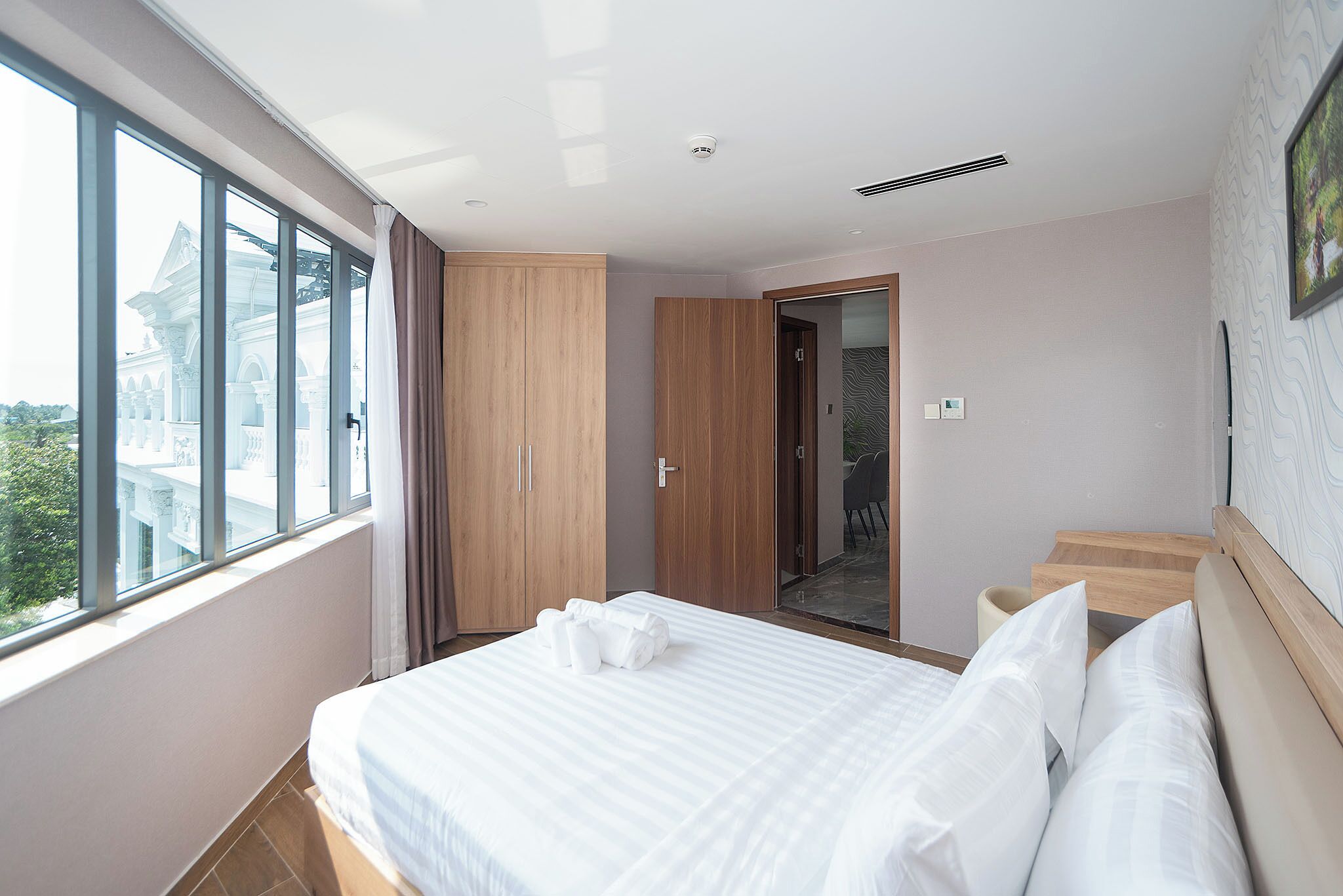 A hotel room with a large window, a bed with white linens, a wooden wardrobe, and a door leading to a balcony.