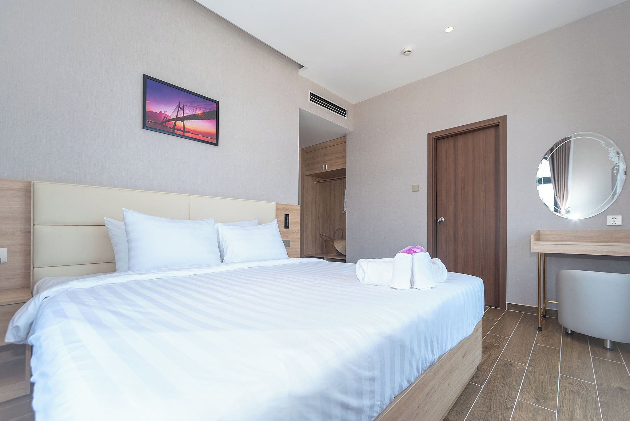 A hotel room with a large bed, a wooden headboard, a bedside table, a mirror, and a picture of a sunset bridge on the wall.