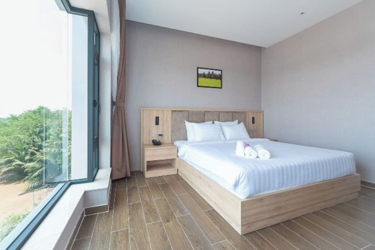 A modern bedroom with a large bed, a nightstand, a window with a view of greenery, and a small picture on the wall.