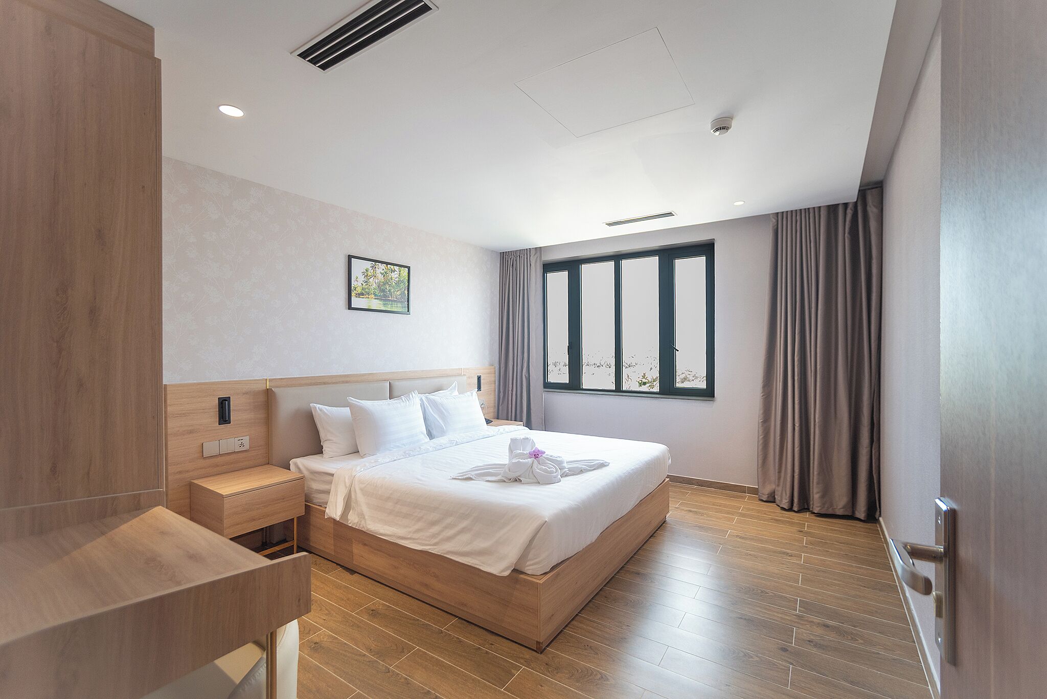 Une chambre d’hôtel moderne avec un lit en bois, une table de chevet, une petite table et une grande fenêtre avec des rideaux.