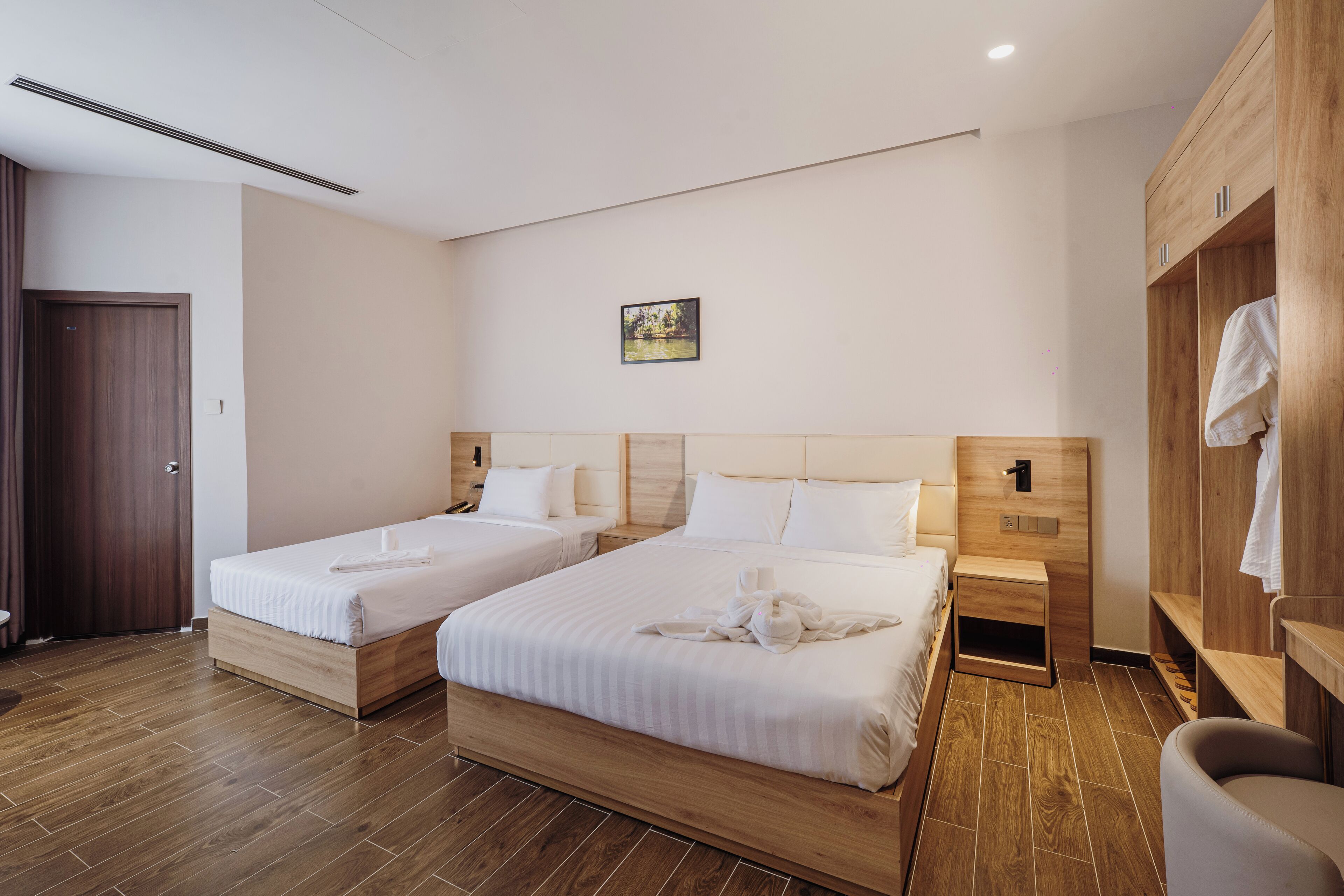 A hotel room with two beds, wooden headboards, a small bedside table, a wooden wardrobe, and a picture on the wall.