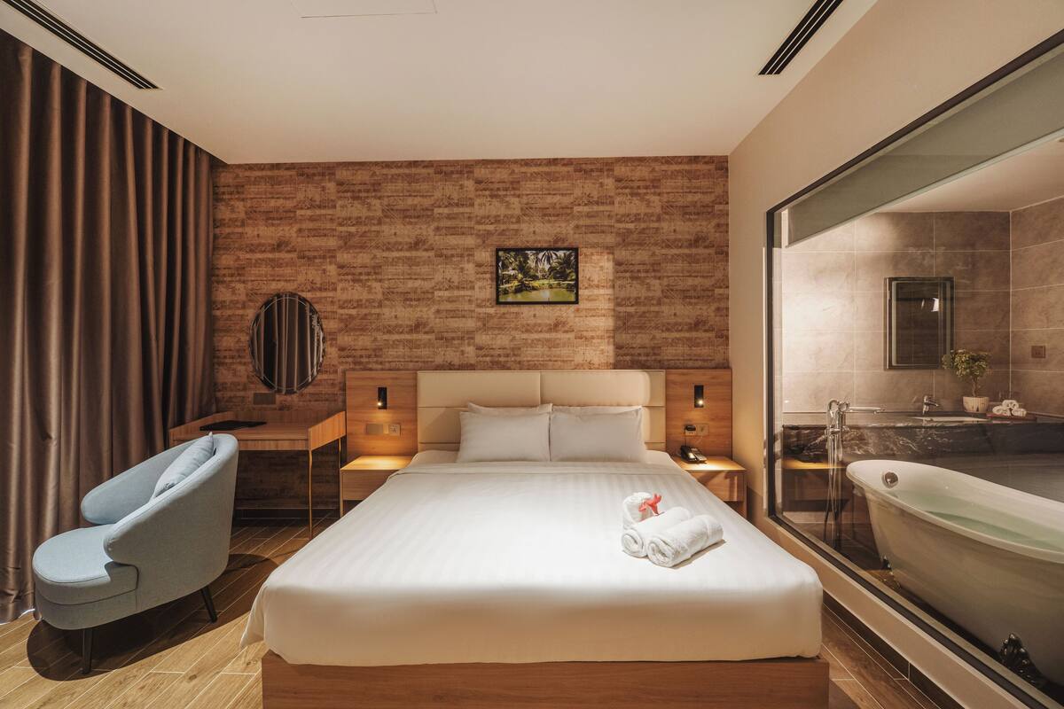 Una moderna camera d'albergo con un letto grande, un'area salotto con una poltrona, un'ampia finestra e un bagno con una vasca.