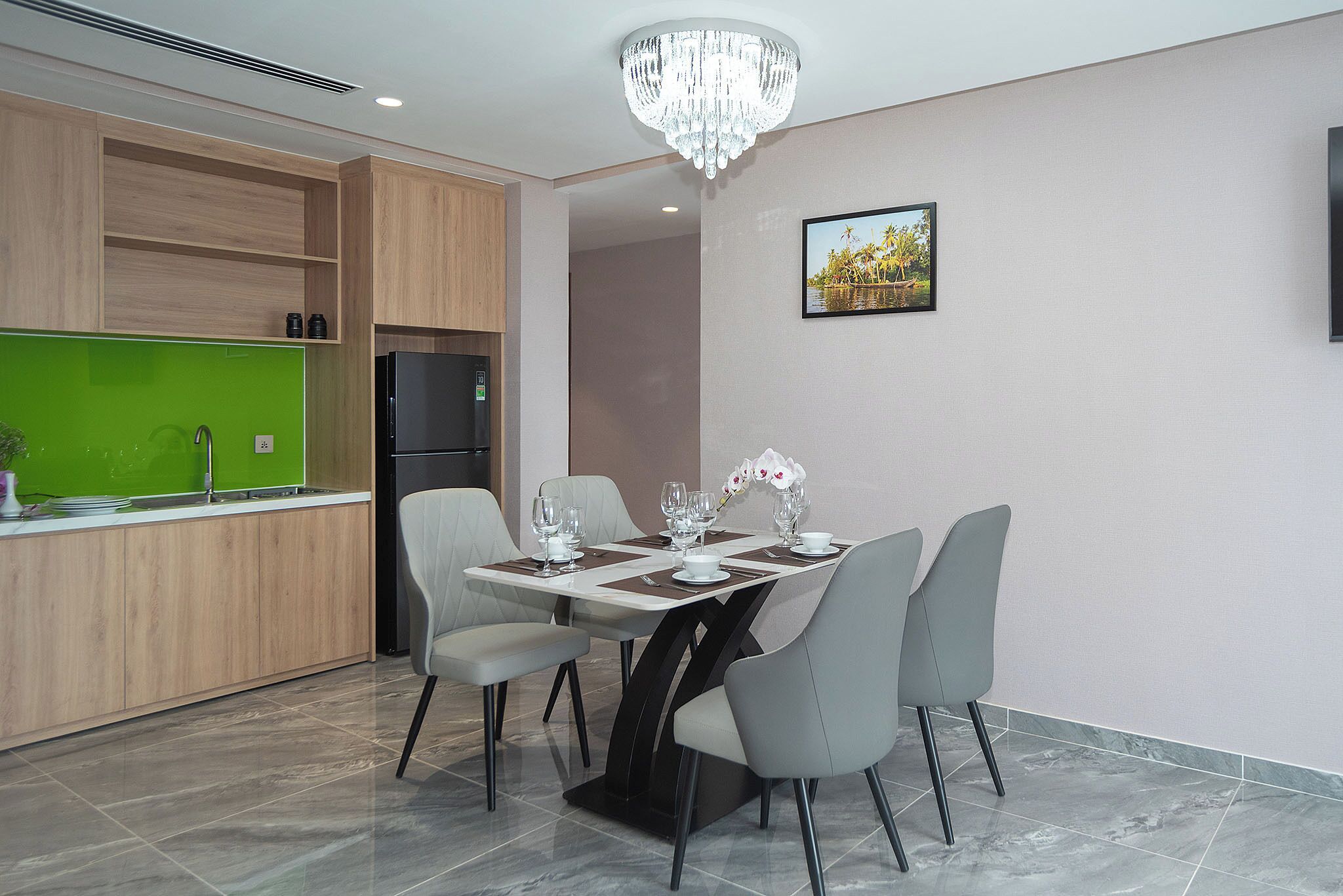 Ein moderner Essbereich mit Marmorfliesen, einem mit weißen Tellern und Gläsern gedeckten Esstisch und grauen Stühlen. Im Hintergrund befindet sich ein schwarzer Kühlschrank und eine grüne Wand.
