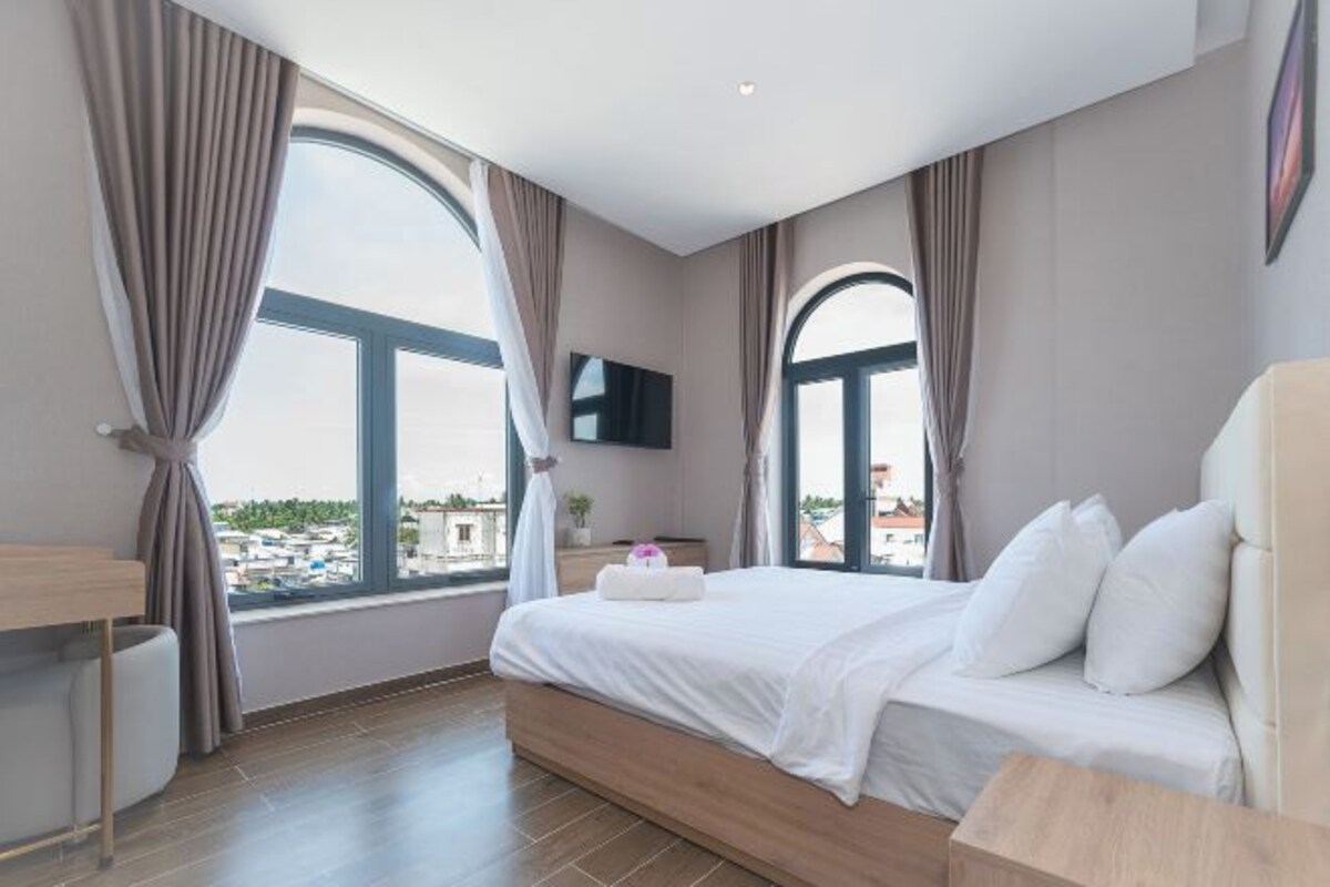 A modern hotel room with a large bed, a TV mounted on the wall, and a view of the city through large windows.