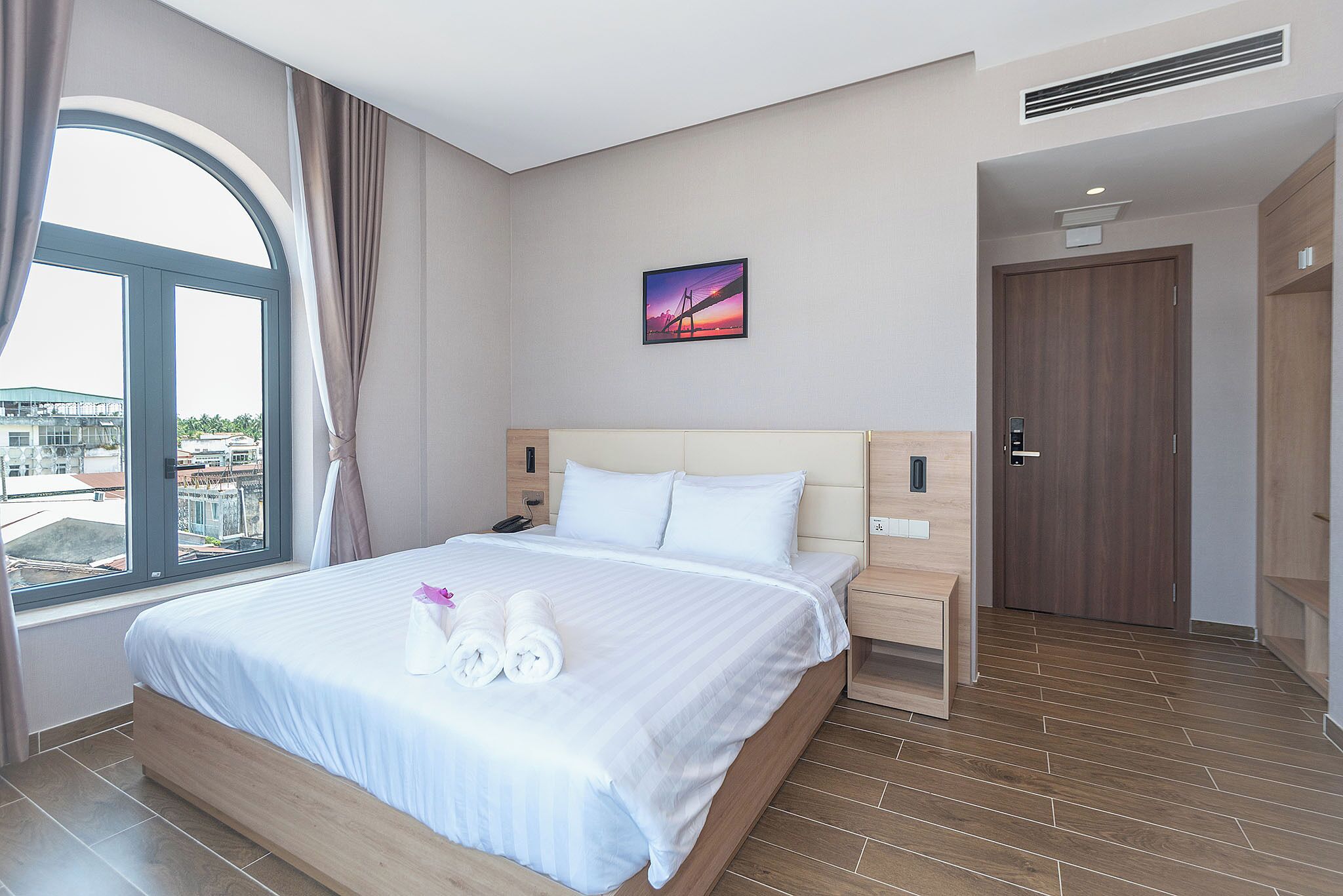A hotel room with a large bed, a bedside table, a nightstand, a window with curtains, a door, and a wall-mounted picture.