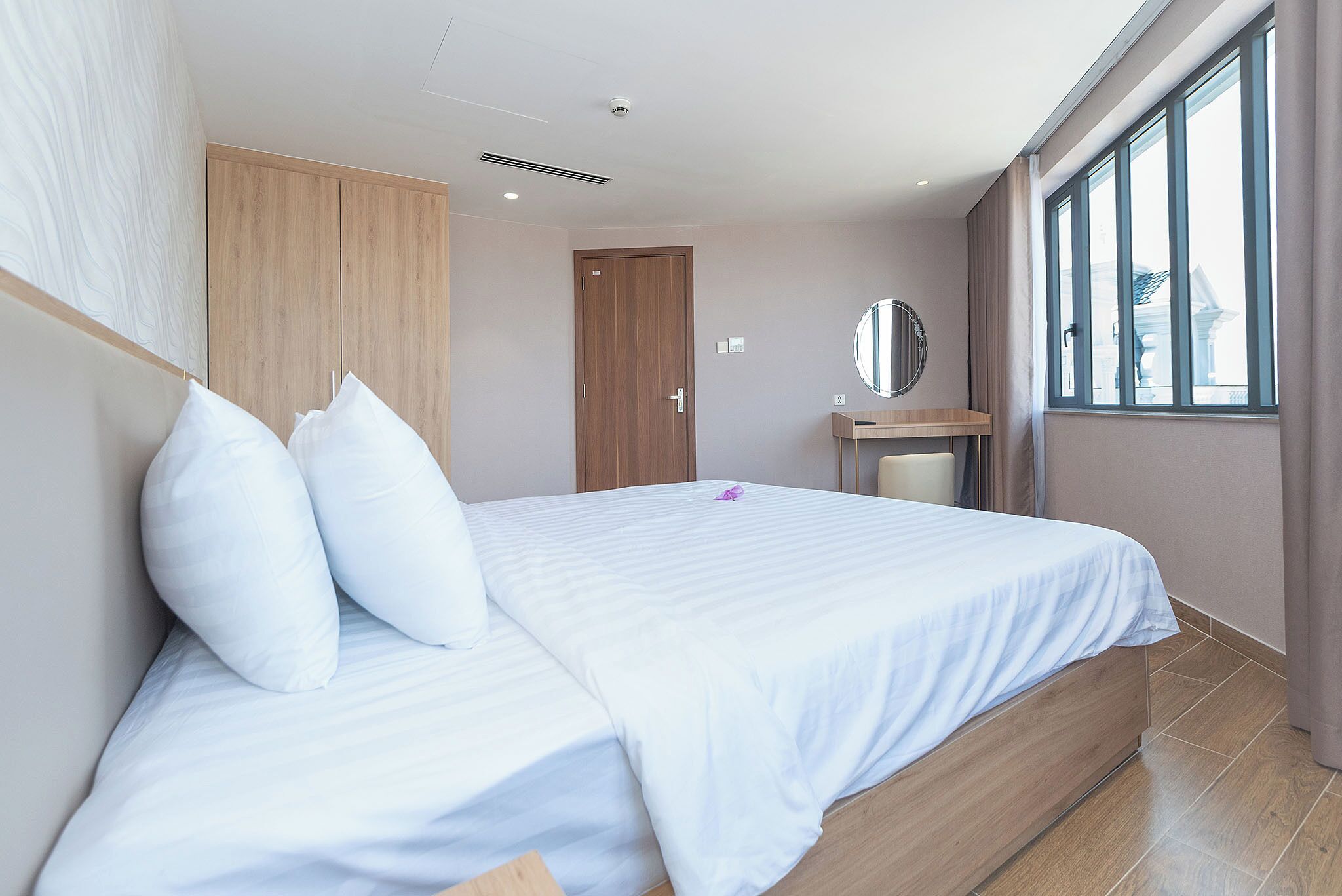 Una habitación de hotel moderna con una cama grande, un armario de madera, un escritorio pequeño y un ventanal amplio.