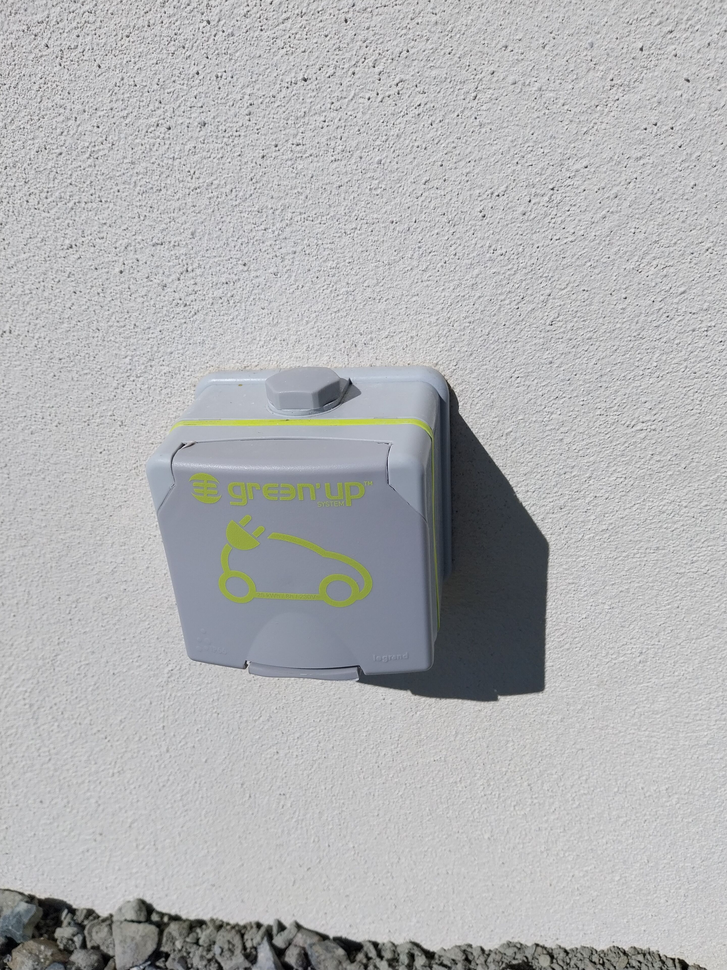 Een grijs, rechthoekig object met de tekst 'Greenup System' en een icoon van een auto, bevestigd aan een witte muur.
