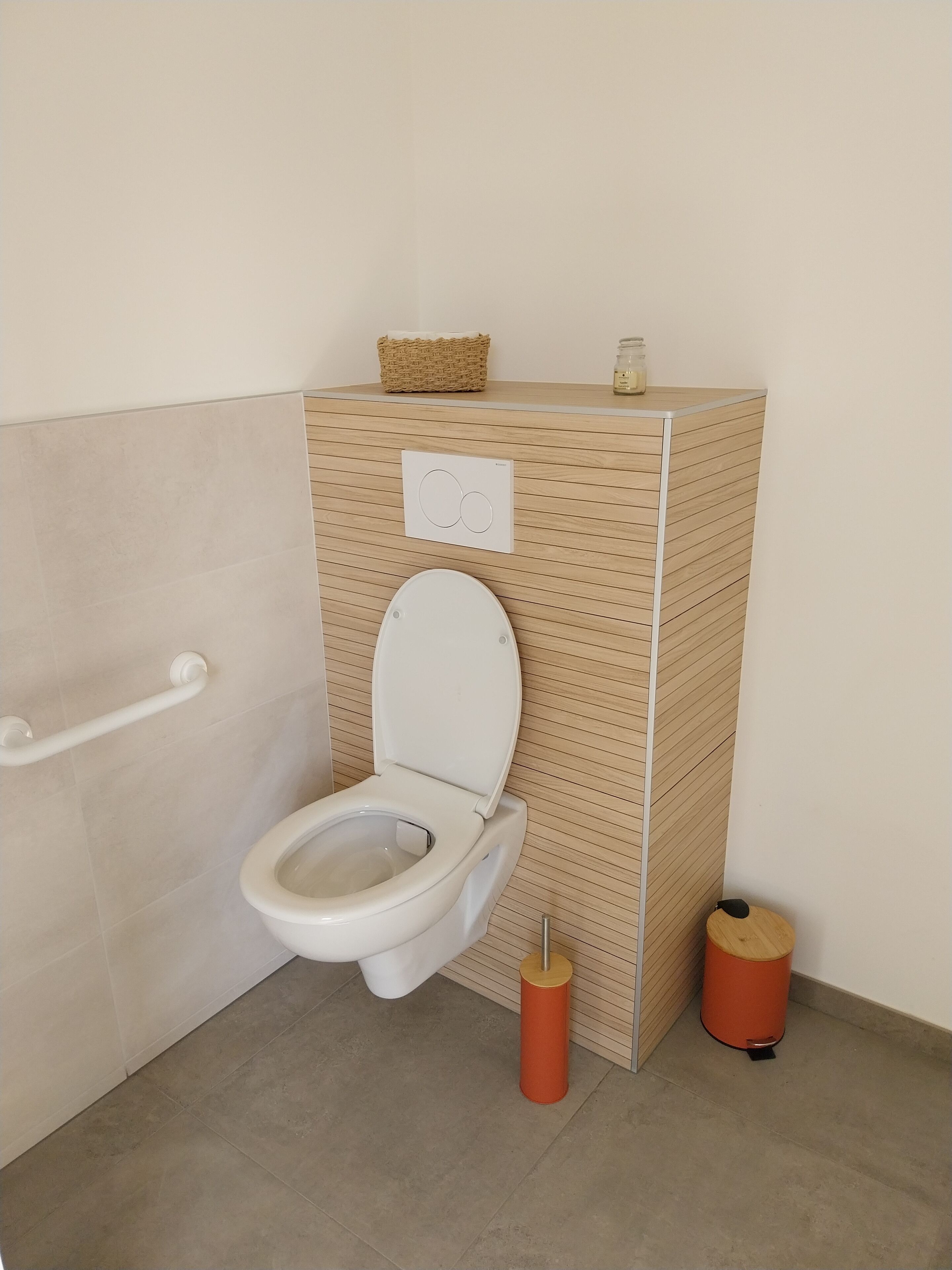 A compact bathroom with a white toilet, a handrail, a waste bin, and a small basket on the wall.