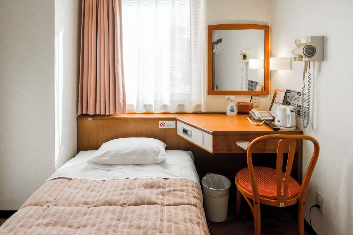 Ett hotellrum med en säng, ett skrivbord med en telefon, en stol, en papperskorg och ett fönster med gardiner.