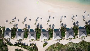 Vista aérea de un complejo turístico en la playa con filas de villas frente al mar, palmeras y una playa de arena.