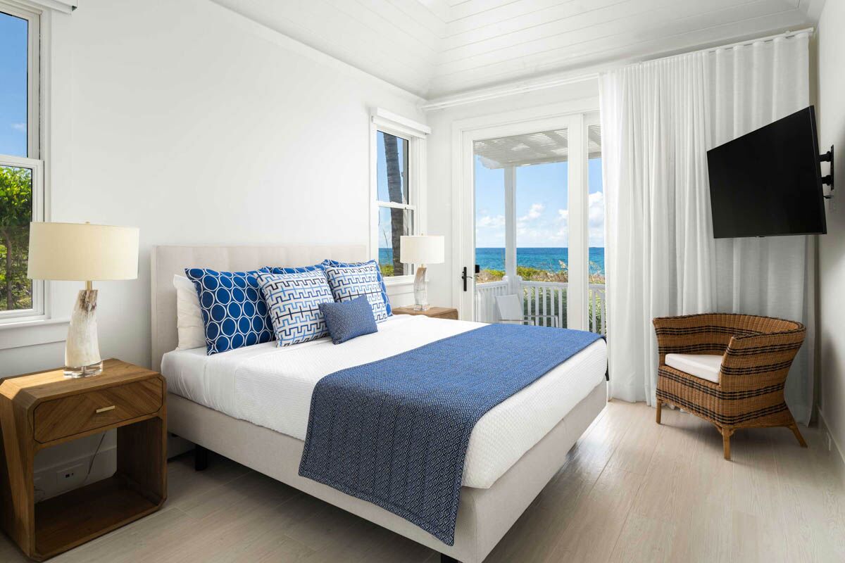 Een slaapkamer met een bed, nachtkastje, rieten stoel, televisie en uitzicht op de oceaan.