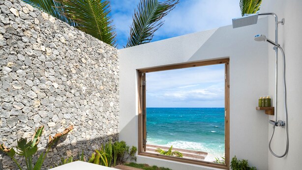 A room with a stone wall, a wooden deck, a white bed, and a view of the ocean. - 4 BR All-Inclusive Villa @ Ambergris Cay: Dream Away (Big Ambergris Cay)