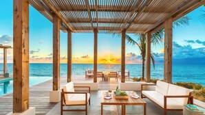 A wooden deck with outdoor seating, overlooking a pool and the ocean at sunset.