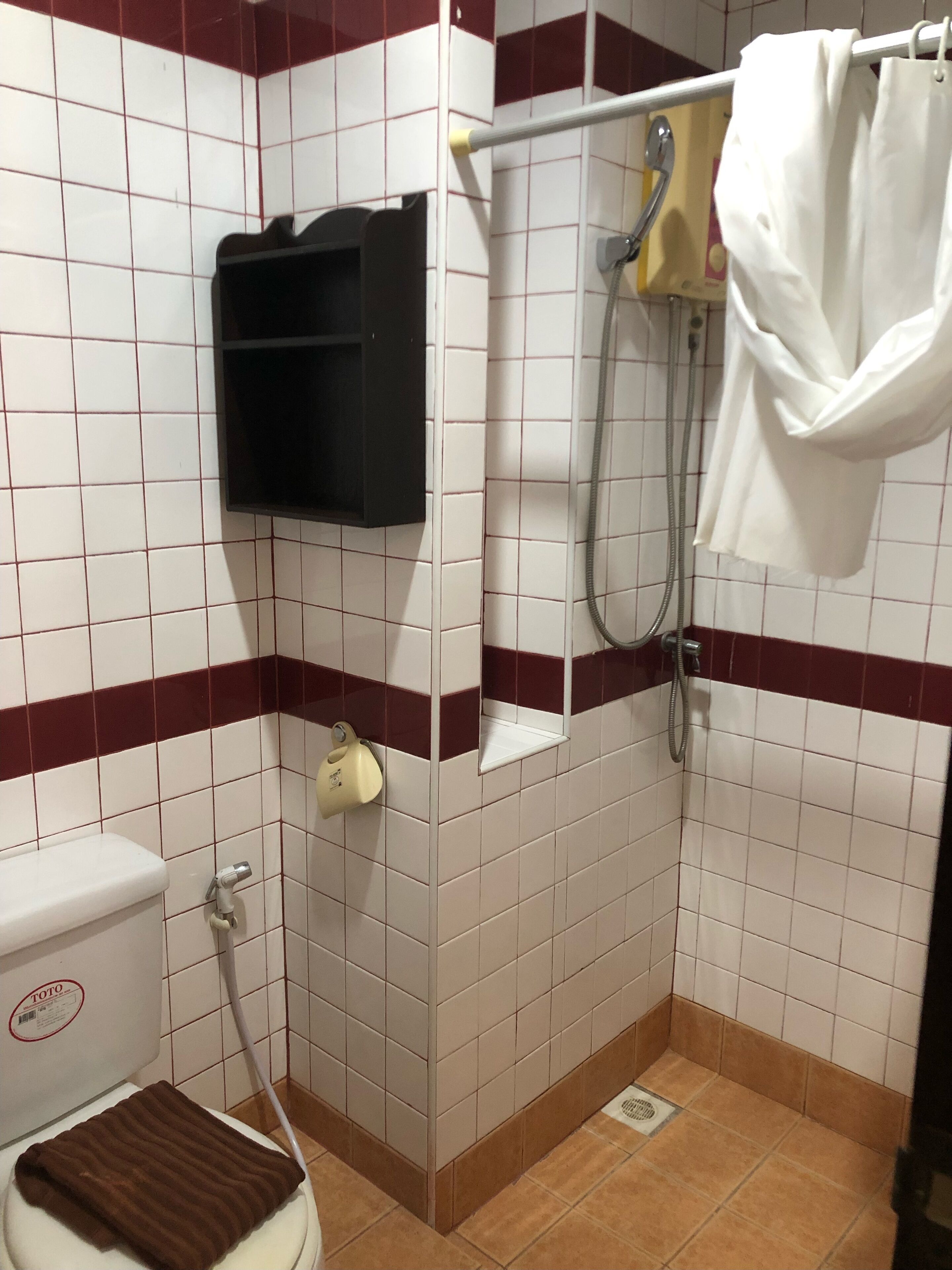 A small bathroom with white and maroon tiles, a toilet, a shower, and a towel rack.