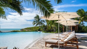 Un espace aménagé au bord de la piscine, avec des chaises longues et des parasols, offrant une vue dégagée sur l’océan, et un décor tropical agrémenté de palmiers.