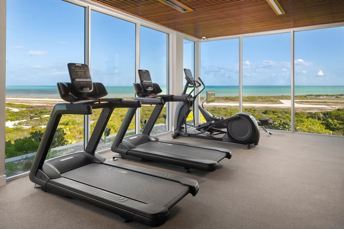 A gym with treadmills and an elliptical machine, large windows with a view of the beach and ocean.