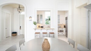 Un comedor amplio con una mesa central y sillas, un jarrón grande y vistas a una cocina con armarios blancos y un ventanal.