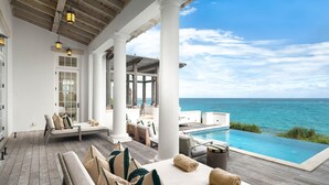 Une piscine à débordement offrant une vue sur l’océan, un espace salon couvert avec des coussins et une terrasse en bois.