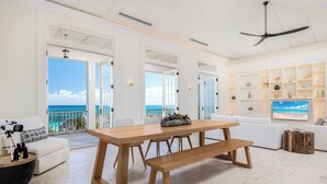 A spacious living room with a wooden dining table, white sofas, and a balcony with a view of the ocean.
