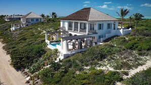 A large white house with a pool, surrounded by lush greenery and a beach.