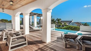 A covered outdoor area with wooden flooring, lounge chairs, and a pool.