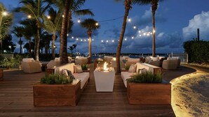 Un espace extérieur aménagé avec des jardinières en bois, des canapés et un foyer central, le tout entouré de palmiers et illuminé de guirlandes lumineuses.