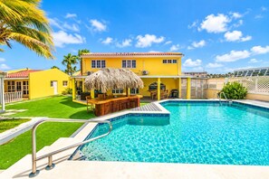 Ein Pool mit Holzterrasse und Bar, umgeben von einem gelben Gebäude mit Strohdach.