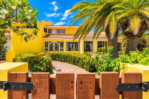 A yellow building with a tiled roof, surrounded by a garden with palm trees and flowering plants.