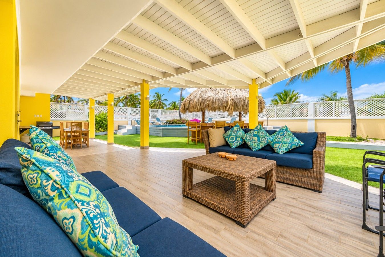 An outdoor lounge area with a covered roof, wicker furniture, and a pool in the background.
