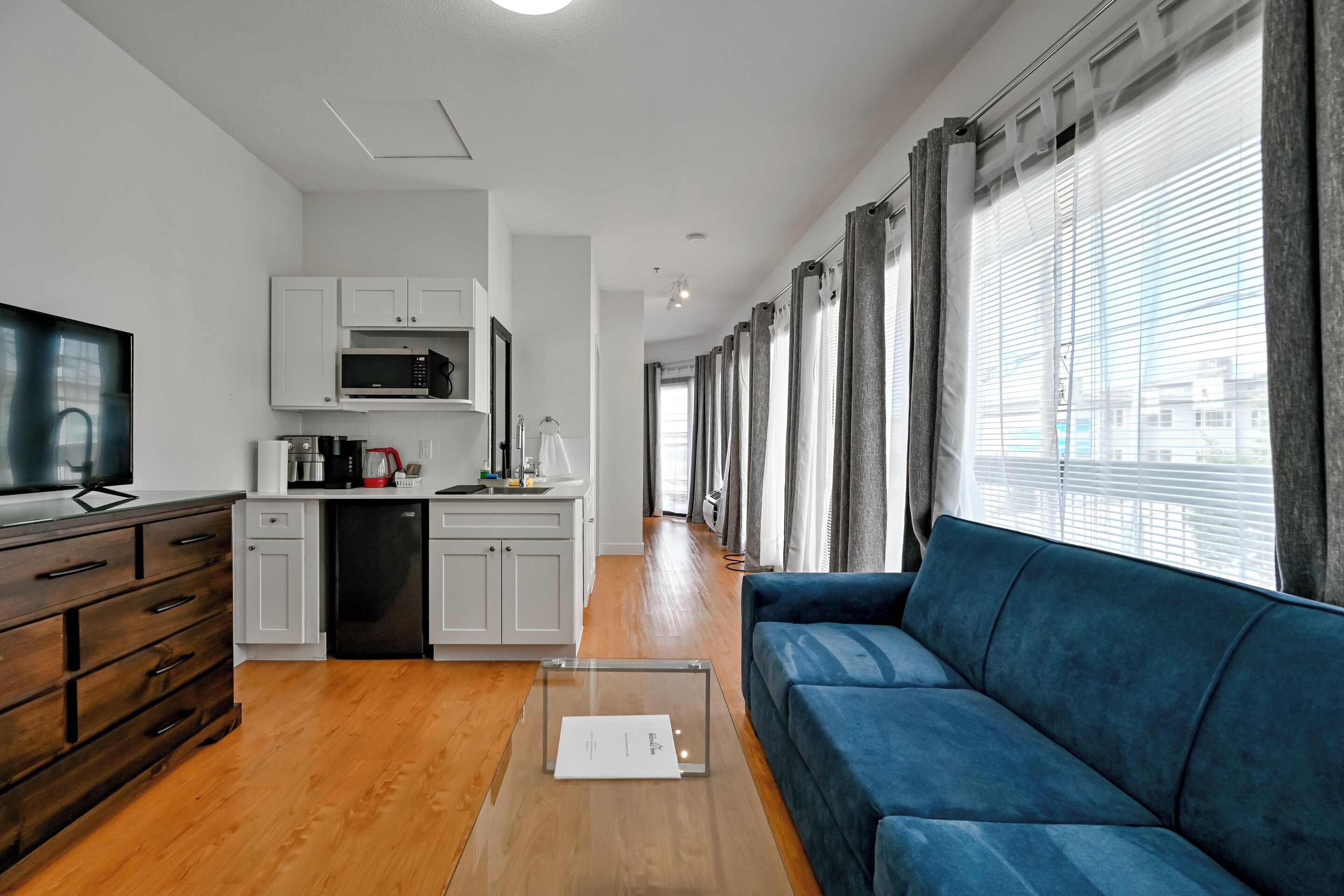 A modern living room with a blue sofa, wooden cabinets, a kitchen area with white cabinets, and a large window with curtains.