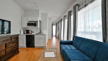 Een moderne woonkamer met een blauwe bank, houten kasten, een keuken met witte kasten en een groot raam met gordijnen.
