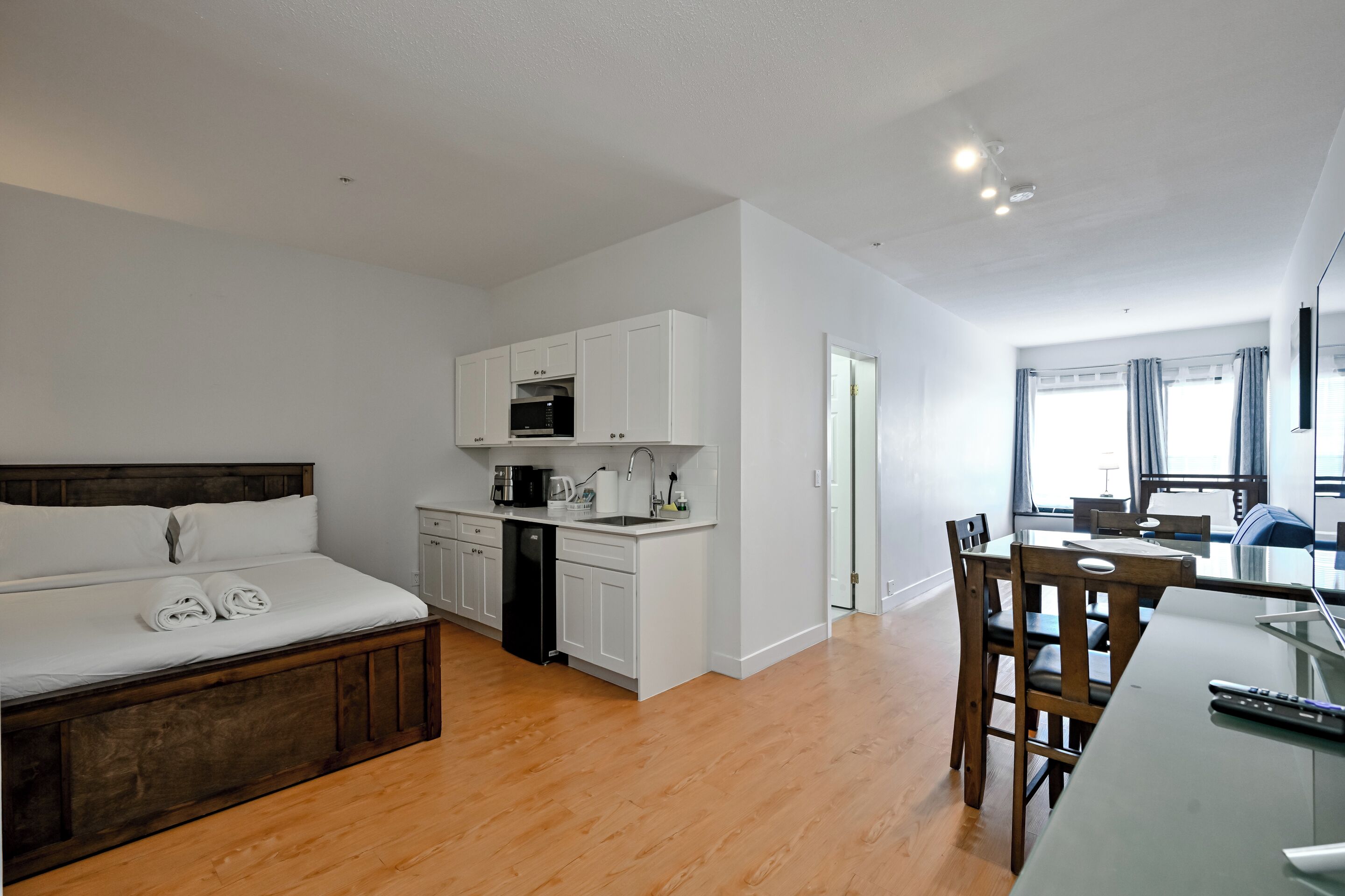 A modern hotel room with a bed, a kitchenette, a dining area, and a view of the city.