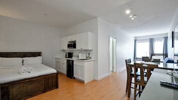 A modern hotel room with a bed, a kitchenette, a dining area, and a view of the city.