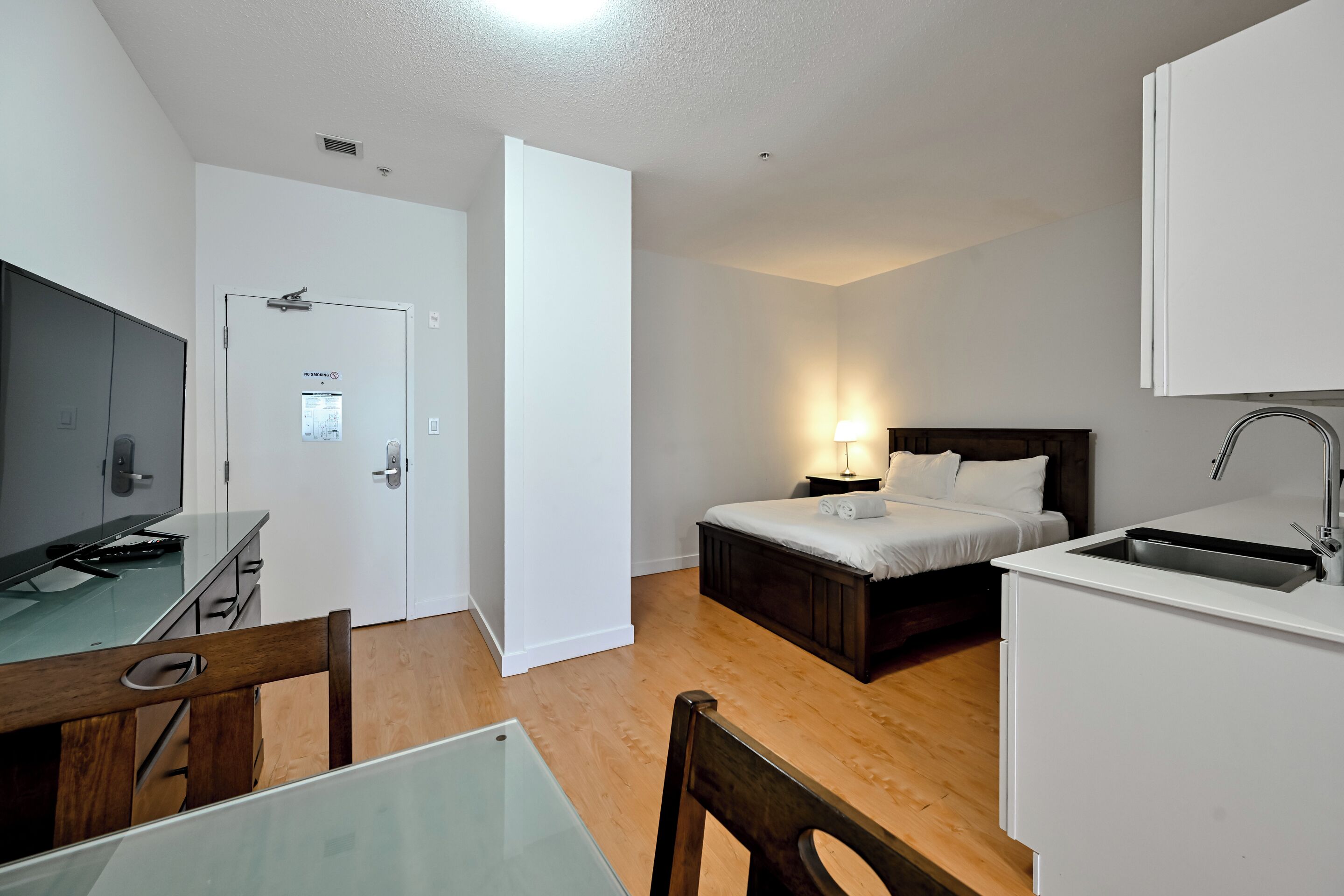 A compact hotel room with a bed, bedside table, desk, chair, and a kitchenette.