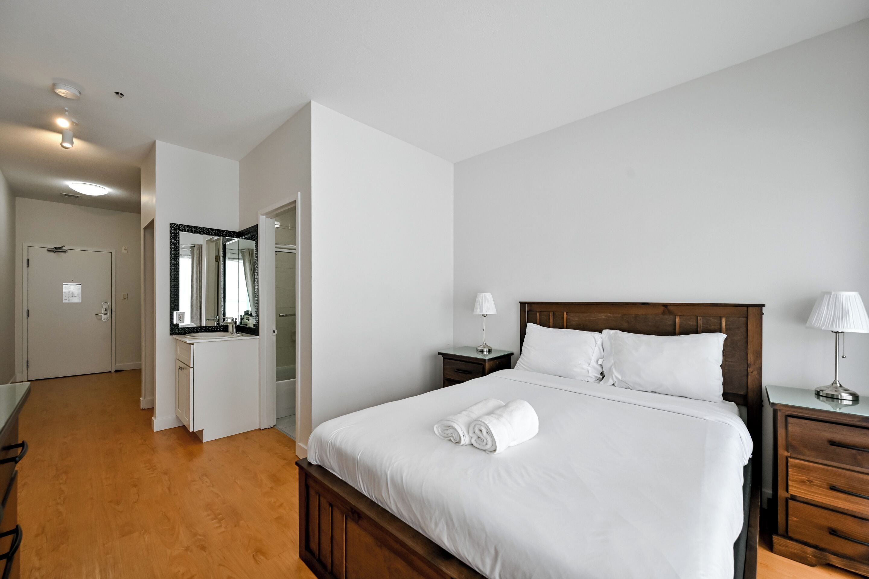 Quarto de hotel com cama de madeira, roupa de cama branca, mesa de cabeceira com abajur, espelho sobre a pia e chão de madeira.
