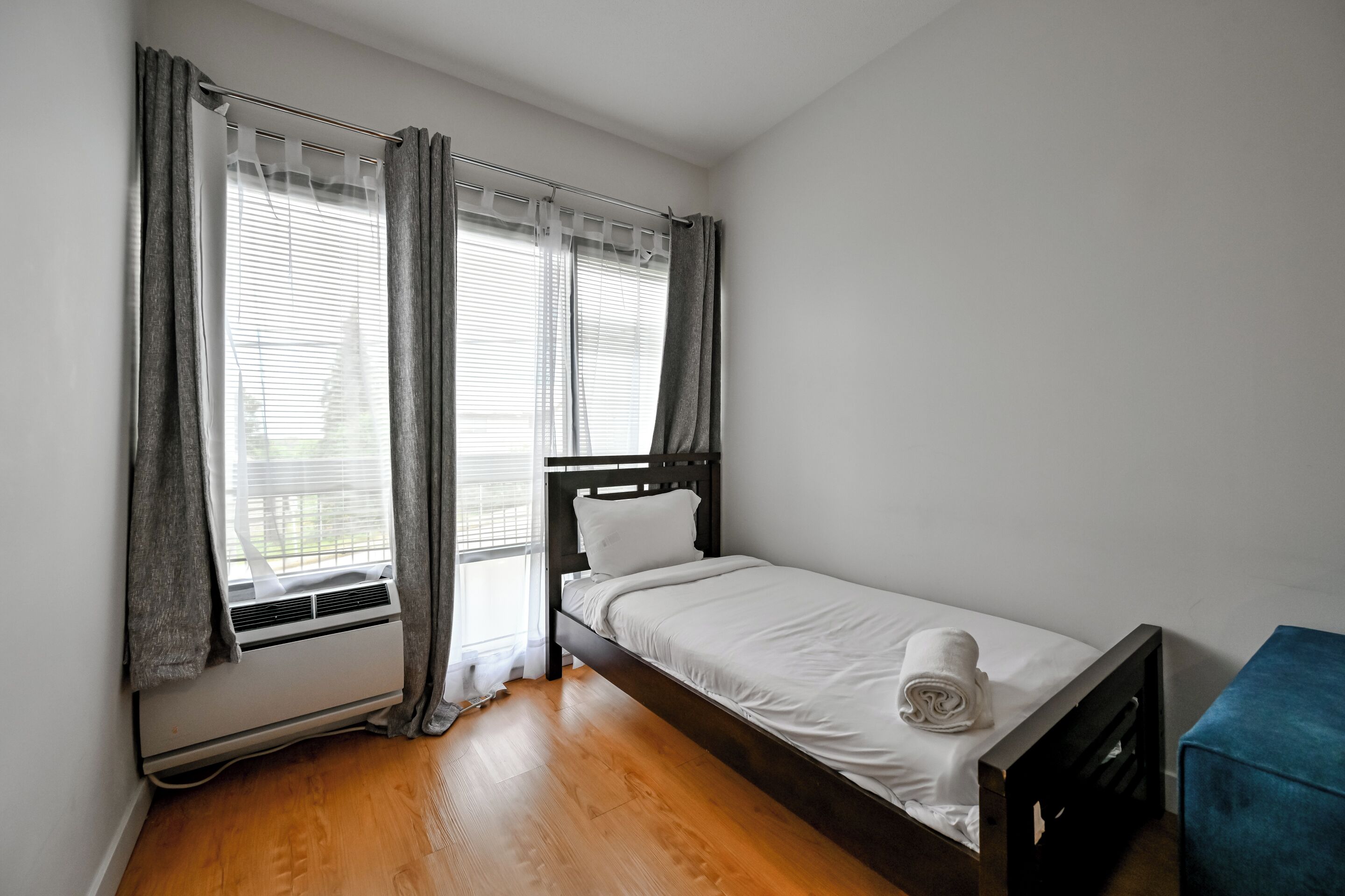 Ein Einzelbett mit Holzrahmen, ein Fenster mit Jalousien, einer Gardinenstange und ein blauer Stuhl.