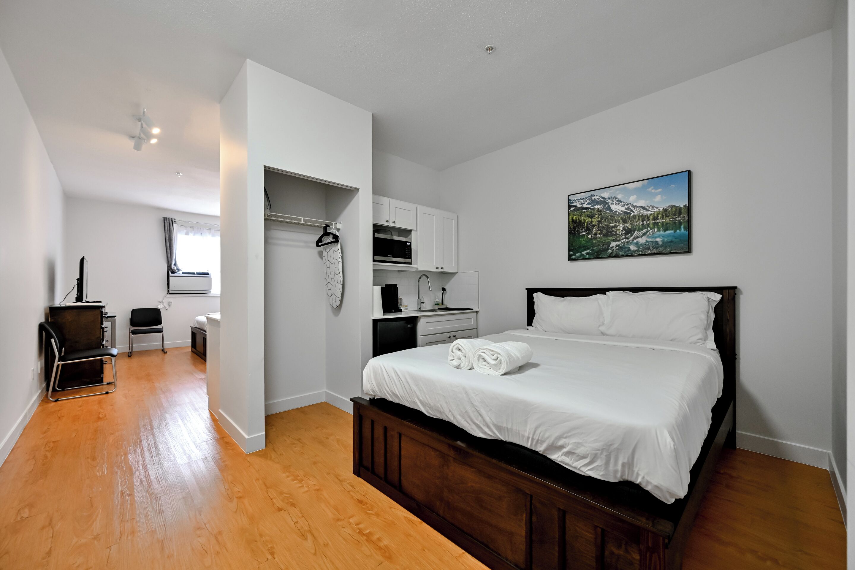 Un dormitorio con cama de madera, ropa de cama blanca, un cuadro de un paisaje montañoso, una kitchenette y una silla.