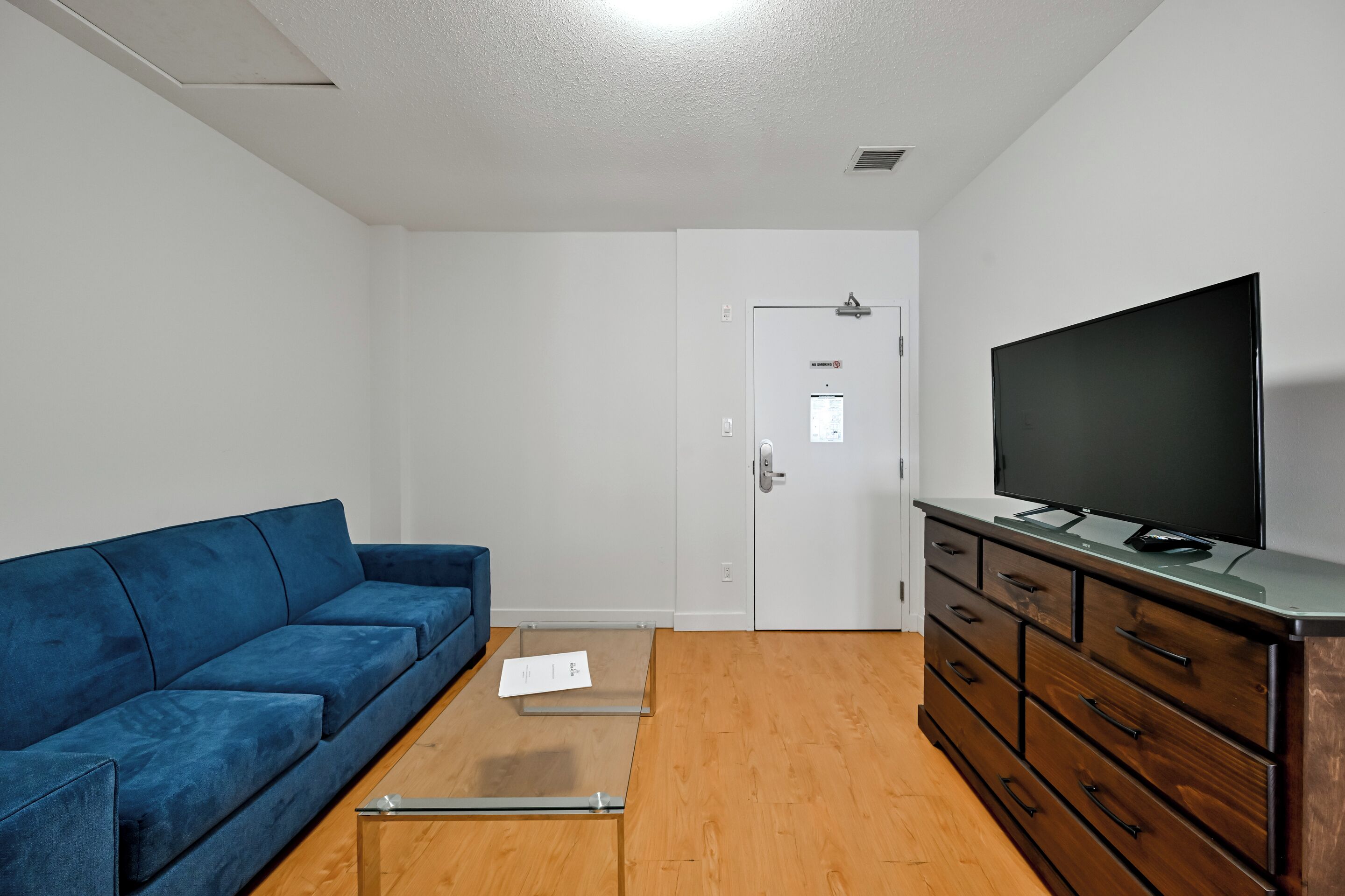 A room with a blue sofa, a TV on a wooden cabinet, a door with a sign, and a small table.