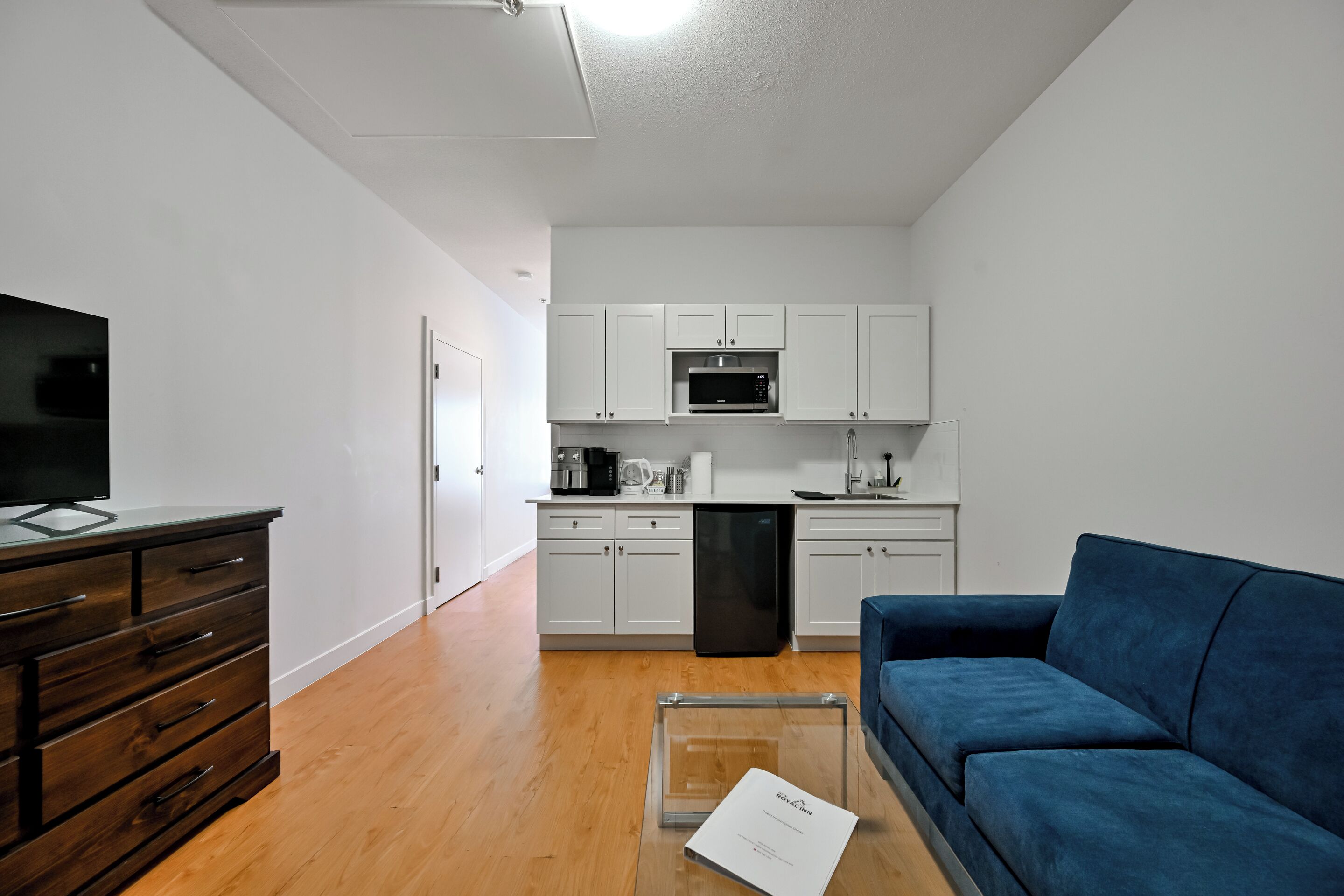 A modern living room with a flat-screen TV, a dark wooden dresser, a blue sofa, and a kitchen area with white cabinets and appliances.