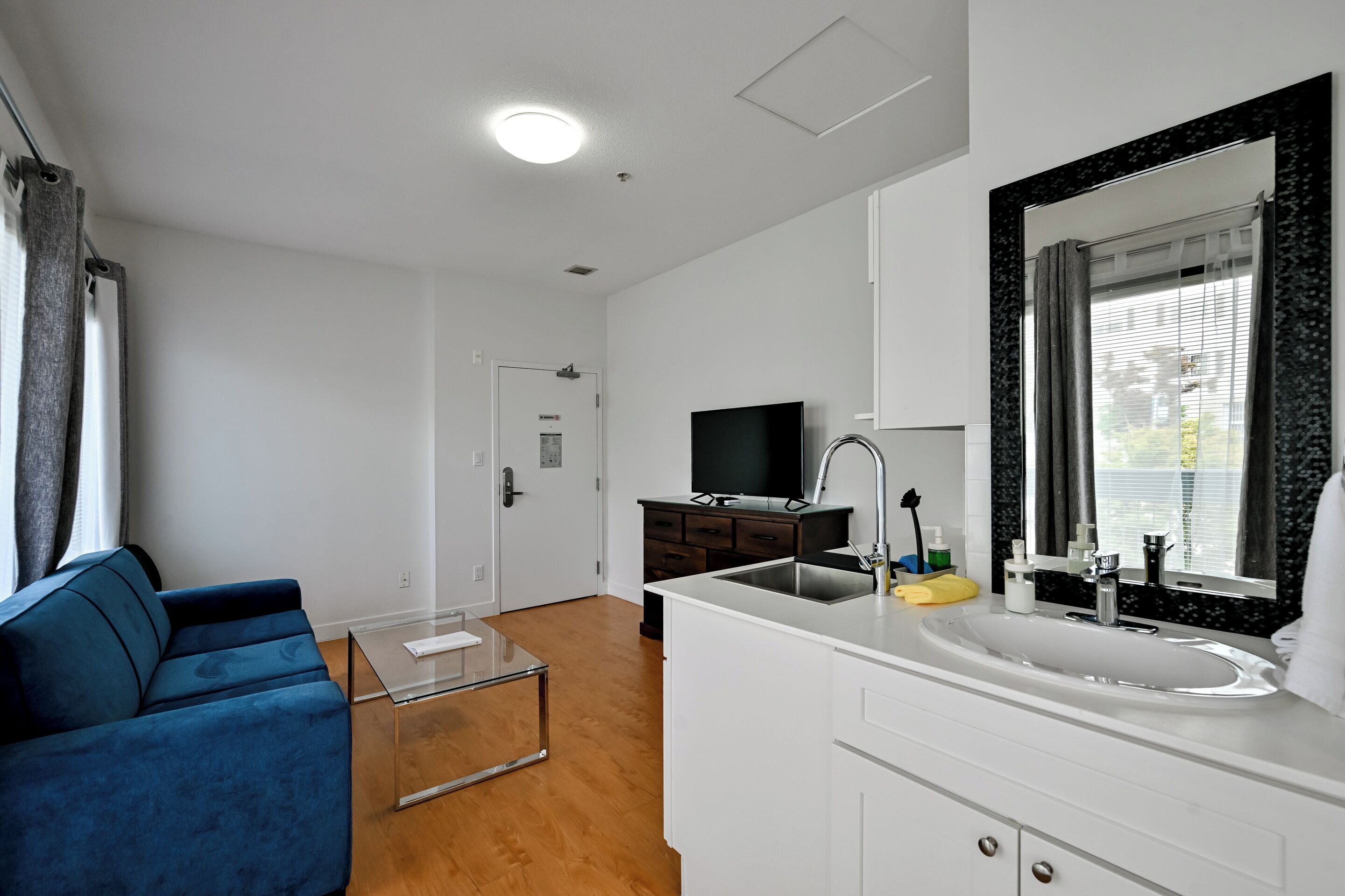 A modern living room with a sofa, a glass coffee table, a TV, a white sink, and a mirror in the kitchen area.