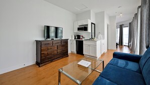 Una sala de estar moderna con una mesa de centro de vidrio, un sofá azul, un aparador de madera oscura, un televisor de pantalla plana y una cocina con armarios blancos y un microondas.