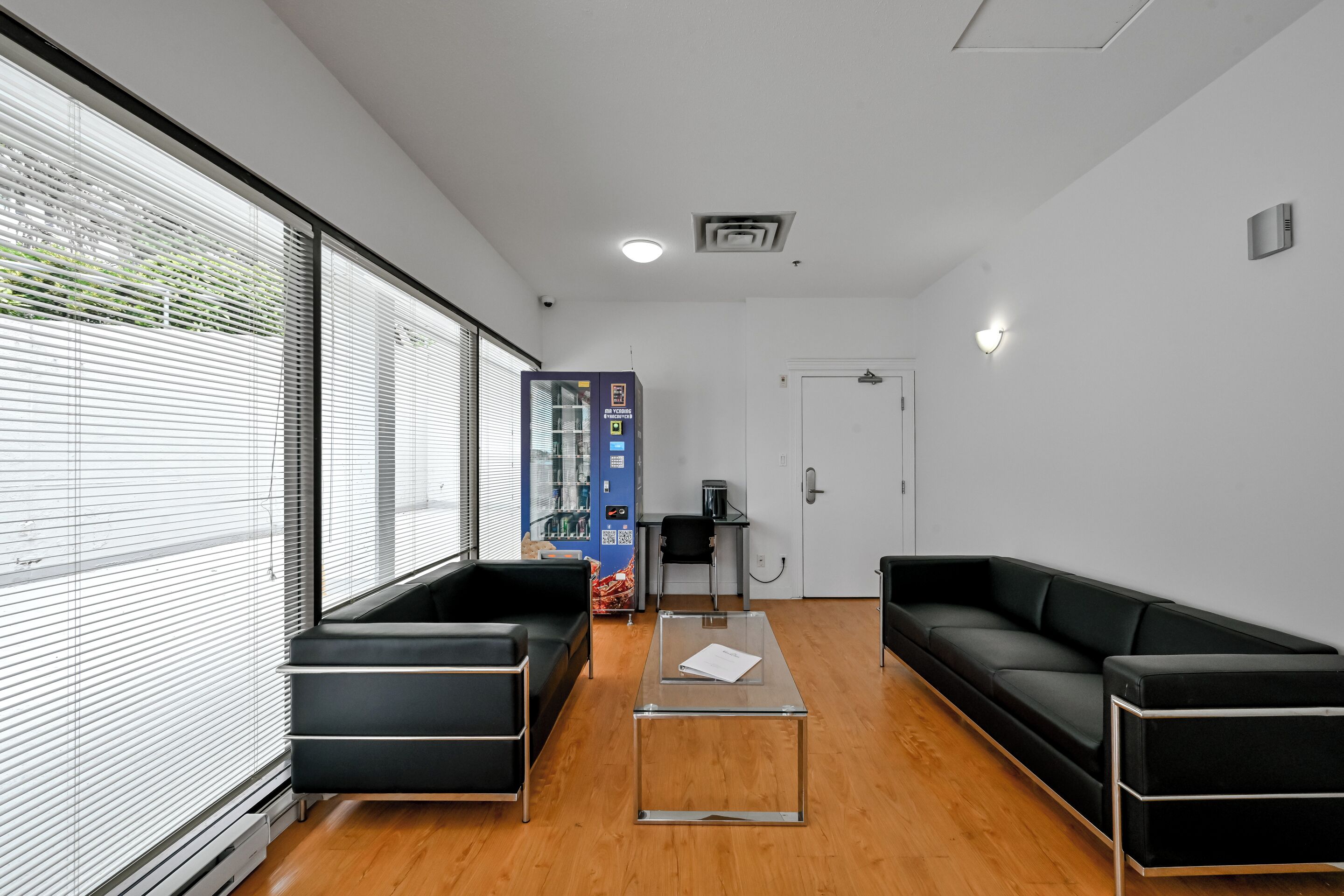 Un área de espera moderna con sofás de cuero negro, una mesa de centro de vidrio y una máquina expendedora.