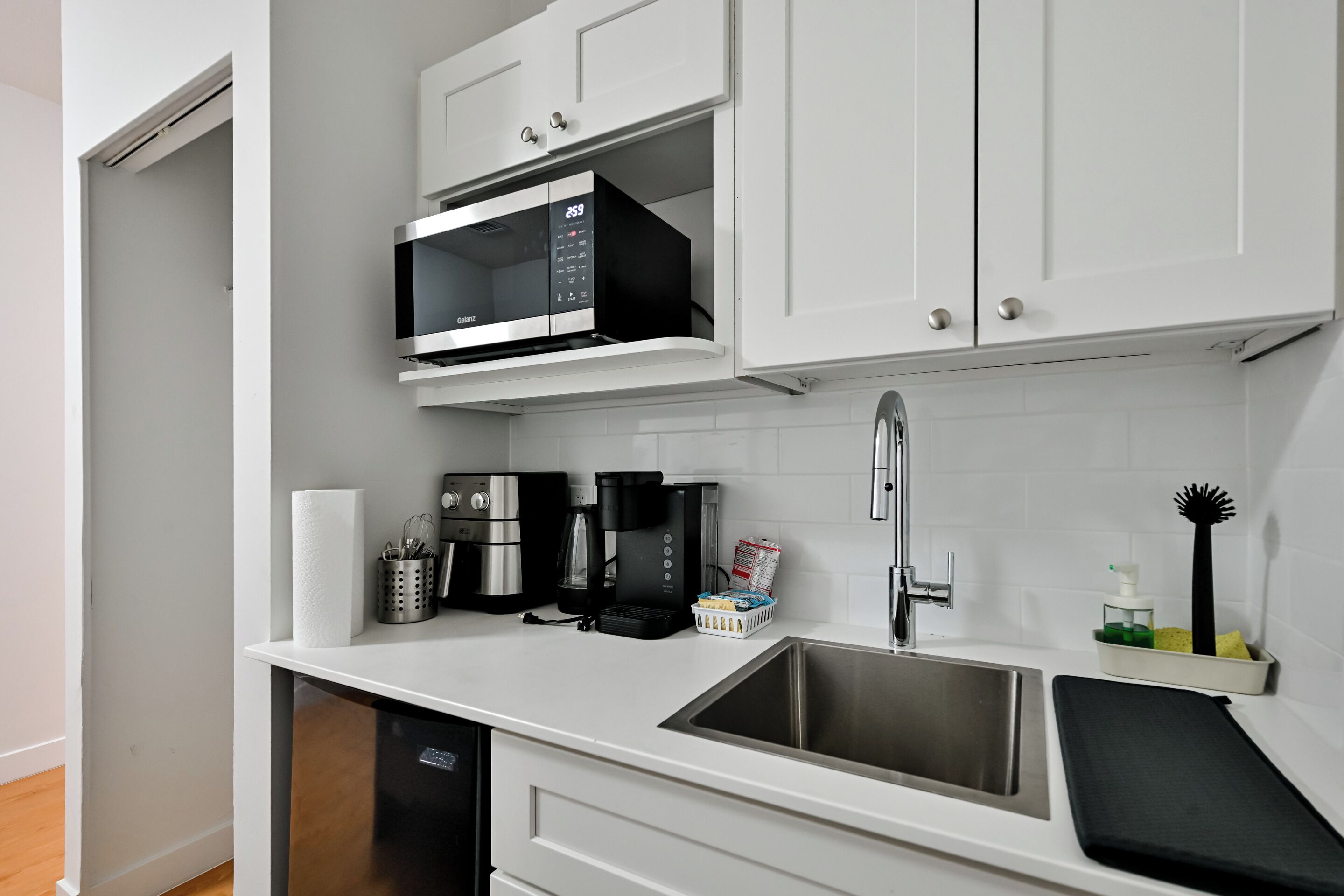 A modern kitchen with white cabinets, a stainless steel sink, a microwave, a coffee maker, and a dish rack.