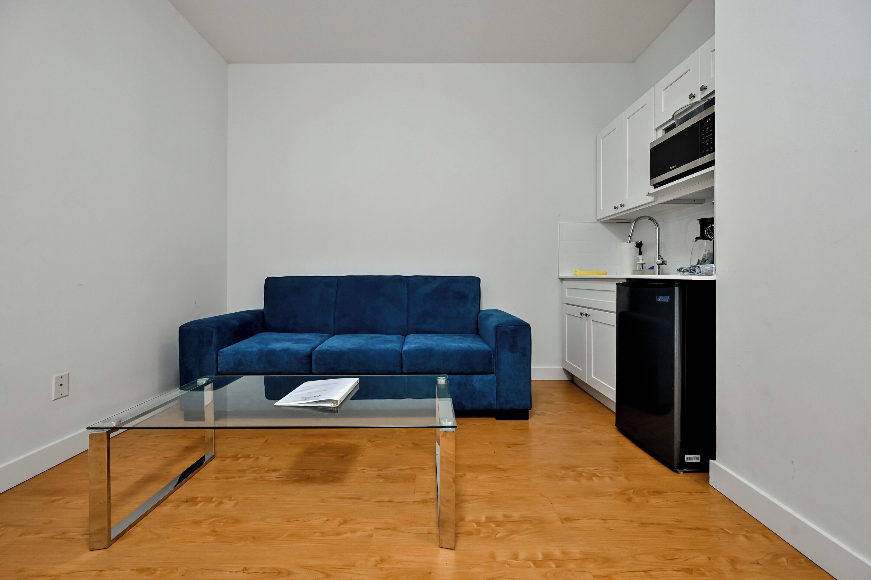 A compact living space with a blue sofa, a glass coffee table, and a kitchenette area.