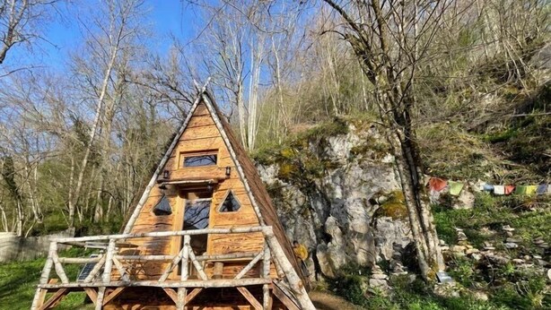 Une cabane en bois avec un toit triangulaire, située sur une pente herbeuse, avec des arbres en arrière-plan.