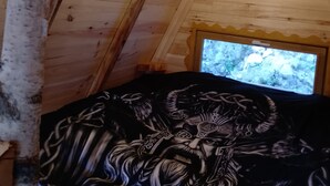 Un intérieur en bois avec un plafond en pente, une fenêtre donnant sur l’extérieur et un lit recouvert d’une couverture à motifs sombres.