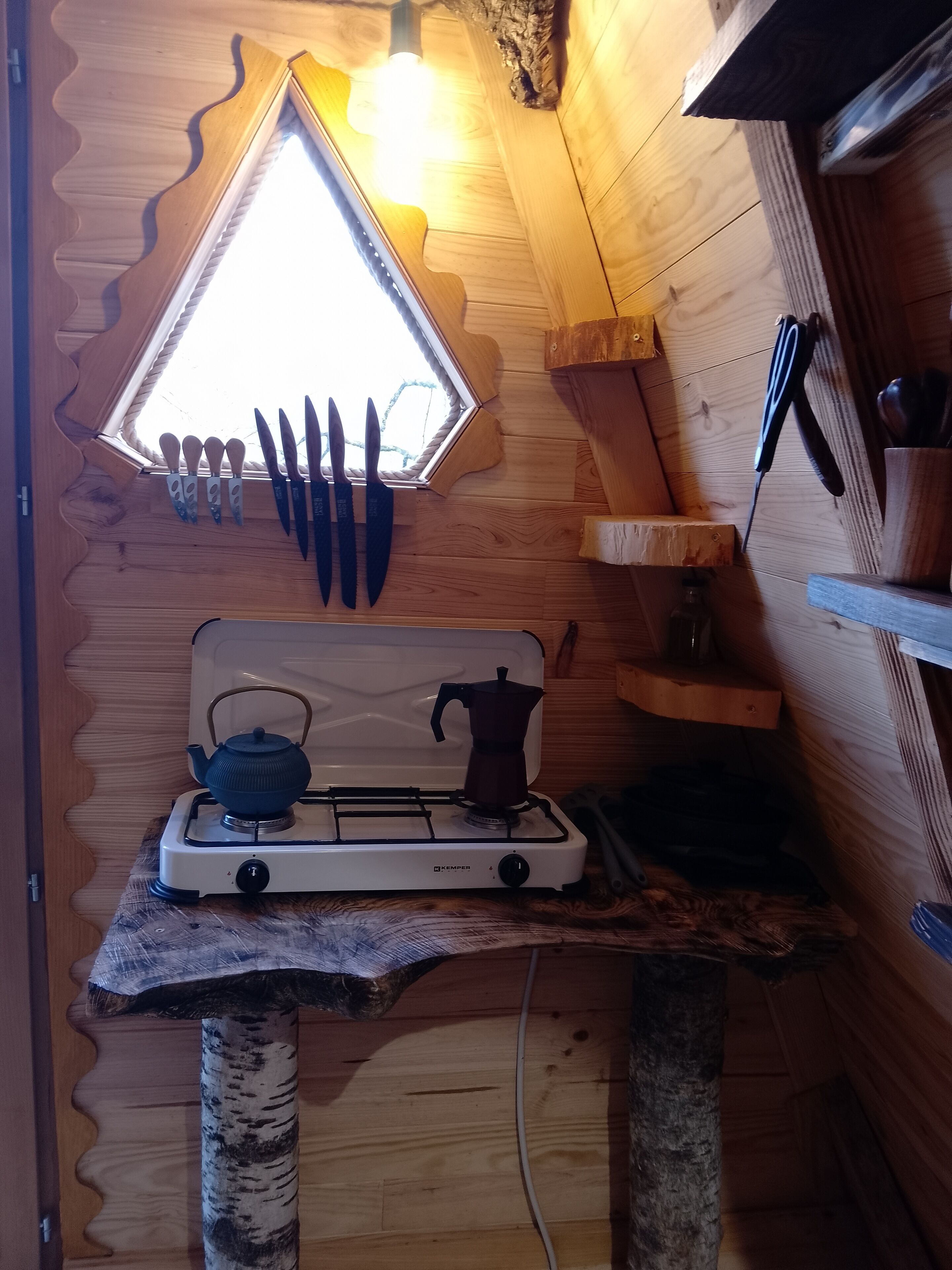 Eine kompakte Küche mit Gasherd, Wasserkocher und Kaffeemaschine. Ein Fenster bietet Blick ins Freie, und ein Satz Messer hängt an der Wand.