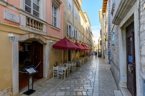 Una estrecha calle adoquinada con mesas al aire libre bajo sombrillas rojas, un letrero y arquitectura europea clásica.